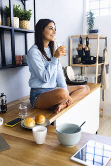 Lächelnde Frau, die im Schneidersitz auf dem Küchentisch sitzt und Saft trinkt - GIOF13597