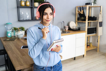 Woman listening music through headphones while holding note pad in kitchen - GIOF13603