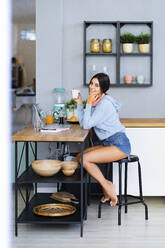 Smiling woman sitting with hand on chin having coffee in kitchen at home - GIOF13606