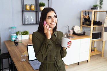 Lächelnde weibliche Fachkraft mit Kaffeetasse, die zu Hause mit einem Mobiltelefon spricht - GIOF13625