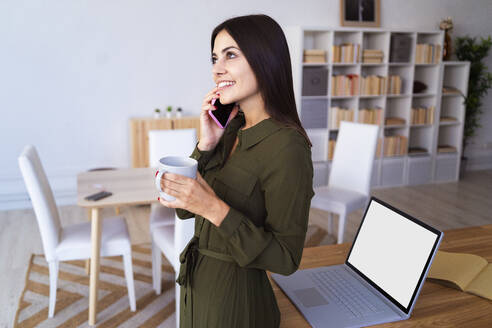 Junge Geschäftsfrau, die zu Hause Kaffee trinkt und mit ihrem Smartphone telefoniert - GIOF13626