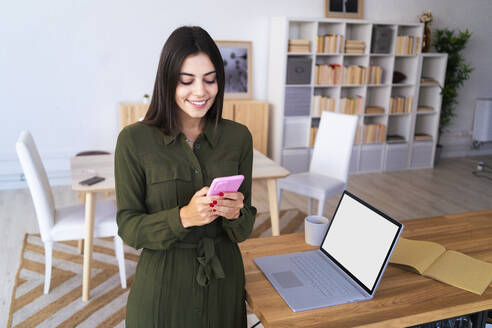 Schöne weibliche Freiberuflerin Text-Messaging über Handy von Laptop zu Hause - GIOF13627