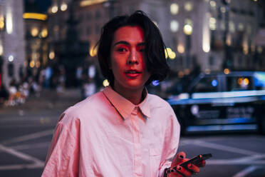 Handsome young man with long black hair holding mobile phone in city at night - AMWF00016
