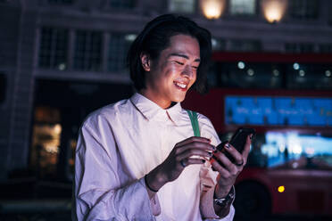 Lächelnder gut aussehender Mann, der nachts in der Stadt mit seinem Mobiltelefon im Internet surft - AMWF00021