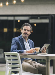 Male professional with digital tablet having coffee at table - JCCMF03985