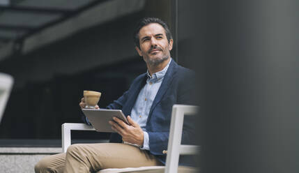 Reifer männlicher Profi, der ein digitales Tablet und eine Kaffeetasse hält, während er auf einem Stuhl sitzt - JCCMF03989