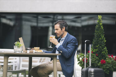Mature businessman drinking juice while working on laptop at hotel - JCCMF03995