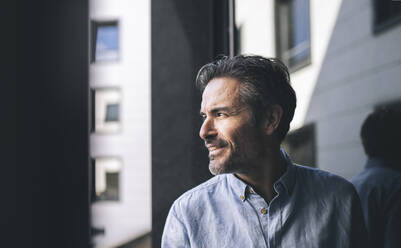 Smiling businessman with hair stubble looking through window - JCCMF04032