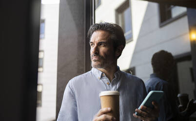 Male freelancer holding mobile phone and disposable coffee cup by window - JCCMF04034