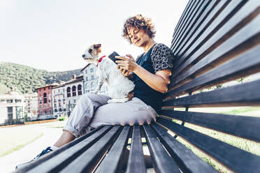 Mid adult woman sitting with pet dog while using smart phone on bench - OMIF00092