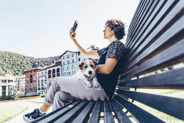 Frau nimmt Selfie durch Smartphone mit Hund auf dem Schoß während sonnigen Tag - OMIF00094