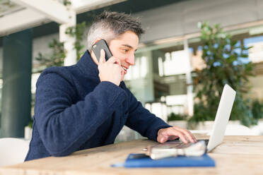 Side view of young male freelancer in casual clothes and eyeglasses talking on mobile phone and looking at camera while sitting at table with laptop and notebook - ADSF30408