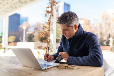 Side view of concentrated young bearded male freelancer in casual clothes and eyeglasses sitting at table with laptop and smartphone while working on project in outdoor cafe - ADSF30409