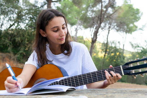 Weiblicher Teenager mit klassischer Gitarre, die einen Akkord spielt, während er Musik in ein Heft im Park schreibt, mit unscharfem Hintergrund - ADSF30425