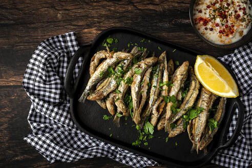 Top view of tasty fried anchovies with juicy lemon piece and chopped parsley on tray against savory sauce - ADSF30428