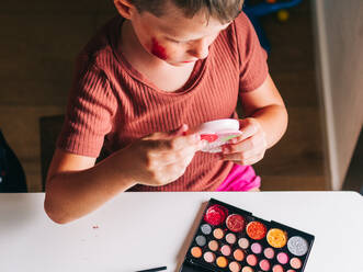 Von oben der Ernte Kind mit Make-up auf Gesicht Blick in den Spiegel am Tisch mit Lidschatten-Palette zu Hause - ADSF30430