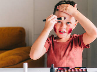 Charmantes Kind mit Make-up-Applikator, das den Kopf berührt, während es in die Kamera schaut, am Tisch mit Lidschattenpalette - ADSF30433