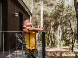 Kleiner Junge steht auf der Veranda eines modernen Ferienhauses im Wald im Sommer - ADSF30438