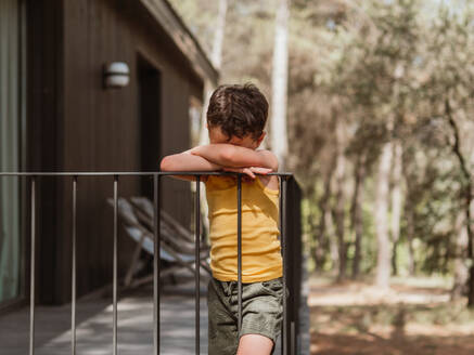 Einsames beleidigtes Kind lehnt an der Holzterrasse eines Hauses im Wald an einem sonnigen Tag - ADSF30439