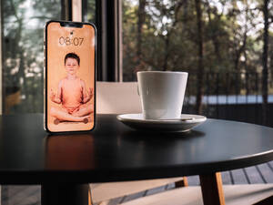 Image of kid doing yoga in Lotus pose on screen of mobile phone placed on table near cup of drink - ADSF30445