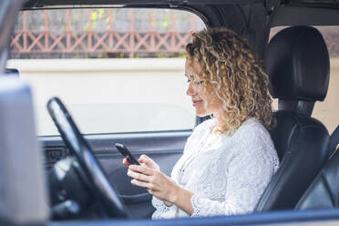 Lächelnde reife blonde Frau, die mit ihrem Smartphone Textnachrichten verschickt, während sie im Auto sitzt - SIPF02410