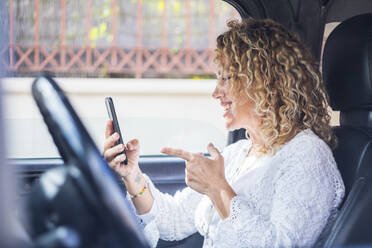 Glückliche Frau mit lockigem Haar gestikuliert während eines Videoanrufs über ein Mobiltelefon im Auto - SIPF02411