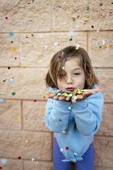 Girl with brown hair blowing colorful confetti in front of wall - RCPF01296