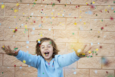 Happy girl with eyes closed throwing confetti in front of wall - RCPF01298