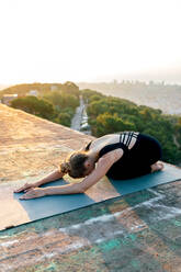 Hoher Winkel Ganzkörper von jungen friedlichen weiblichen Üben Utthita Balasana auf Sportmatte während Yoga-Sitzung auf dem Dach bei Sonnenuntergang - ADSF30487