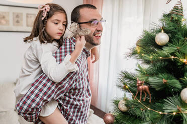 Vater, der seine Tochter Huckepack nimmt, schmückt die Weihnachtstanne mit einem festlichen Stern und bereitet sich auf das Fest vor - ADSF30512