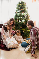Lächelnde Eltern mit Töchtern beim Öffnen von Geschenkkartons auf dem Boden, während sie Neujahr zu Hause feiern - ADSF30515