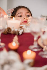Tender child contemplating flaming candle in glass on table with coniferous cones during New Year holiday at home - ADSF30524