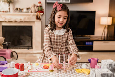 Attentive female child in checkered dress with toy candlestick near chopping board while playing in house - ADSF30527