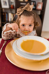 Charmantes Kind mit Schleife auf braunem Haar und Löffel schaut in die Kamera vor einem Teller mit Kürbispüree-Suppe im Haus - ADSF30532