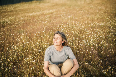 Lächelnde reife Frau, die ihre Knie umarmt, während sie auf einem Feld sitzt - GRCF00959
