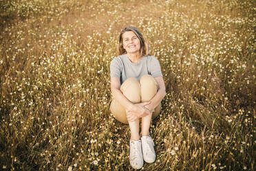 Glückliche Frau, die ihre Knie umarmt, während sie am Wochenende auf einem Feld sitzt - GRCF00961
