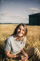 Lächelnde blonde Frau, die Blumen hält, während sie an einem sonnigen Tag in einem landwirtschaftlichen Feld sitzt - GRCF00968