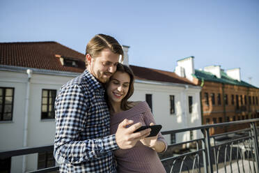 Young couple sharing mobile phone at rooftop - LLUF00106