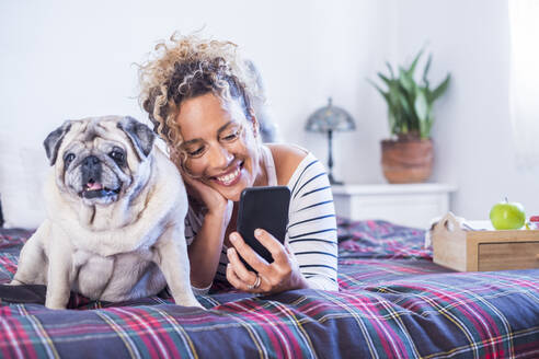 Lächelnde Frau, die ein Mobiltelefon benutzt, während sie mit einem Hund im Schlafzimmer auf dem Bett liegt - SIPF02419