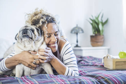 Happy woman embracing pug on bed at home - SIPF02421