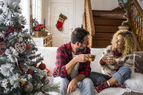Lächelnde Frau, die mit einem Mann Kaffee trinkt, während sie zu Hause auf dem Sofa sitzt, während Weihnachten - SIPF02424