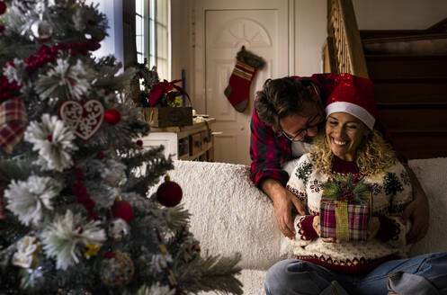 Mann umarmt Frau mit Weihnachtsgeschenk, während er zu Hause auf dem Sofa sitzt - SIPF02427