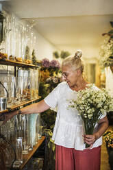 Reife weibliche Floristin mit blühender Pflanze, die eine Vase im Blumenladen auswählt - GRCF00989