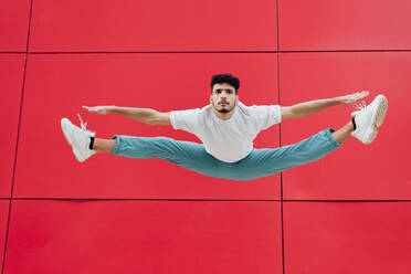 Man with arms outstretched doing splits in front of red wall - MIMFF00722