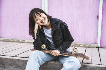 Happy teenager girl with skateboard sitting in front of wall - UUF24720