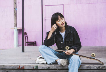 Female teenager with skateboard listening music through in-ear headphones - UUF24724