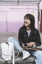 Happy teenage girl with skateboard and backpack sitting on boardwalk - UUF24727