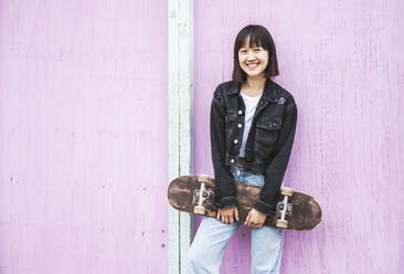 Glückliches Teenager-Mädchen hält Skateboard vor einer Wand - UUF24737