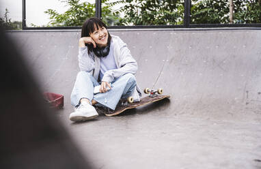 Lächelndes jugendliches Mädchen mit Skateboard im Park sitzend - UUF24738