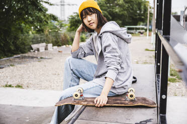 Weiblicher Teenager mit Skateboard auf einer Bank sitzend - UUF24747